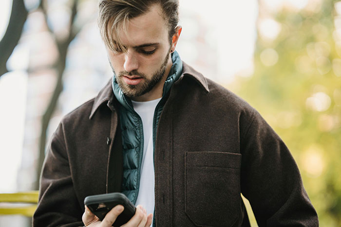 A man standing outdoors, looking at a smartphone, wearing a brown jacket and a blue scarf, during a sunny day.