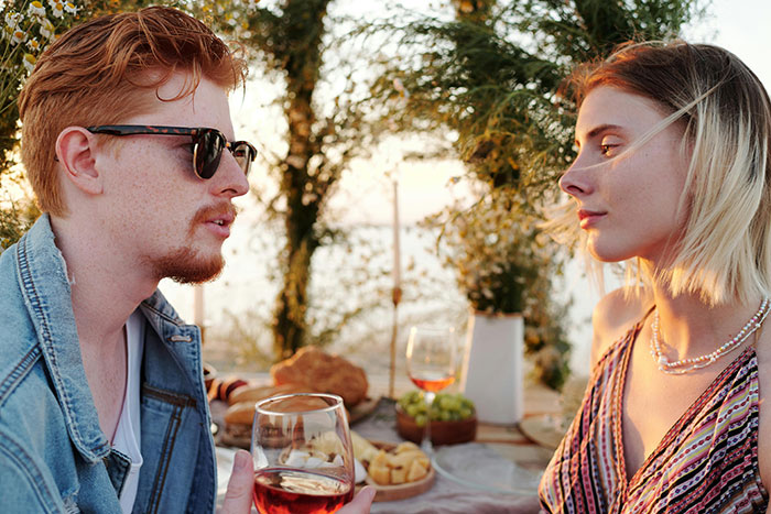 Couple sharing confessions over drinks, hinting at an affair, with a sunset and picnic setting in the background.