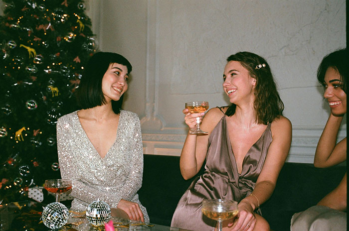 Women at a holiday party sharing drinks, sitting by a Christmas tree, conversing and smiling with a festive ambiance.