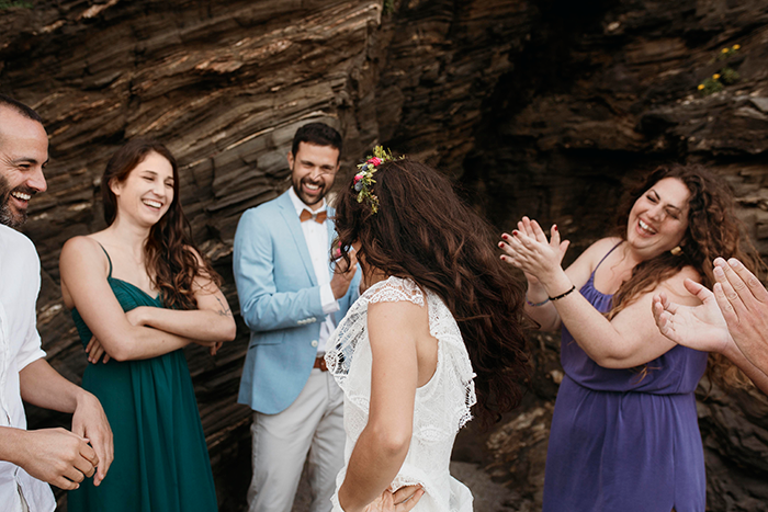 Pregnant bride smiling with friends at wedding, wearing a floral crown, with rocks in the background.