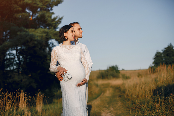 Pregnant bride in a white dress embraces groom outdoors, smiling under clear sky.