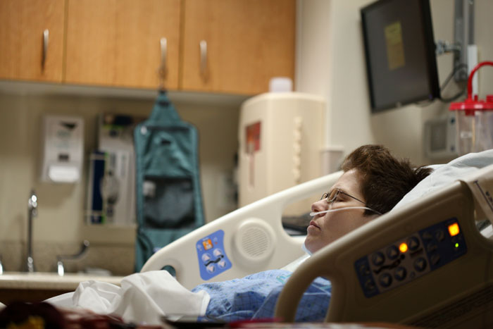 Pregnant woman on life support in hospital bed, connected to medical equipment, in a dimly lit room.