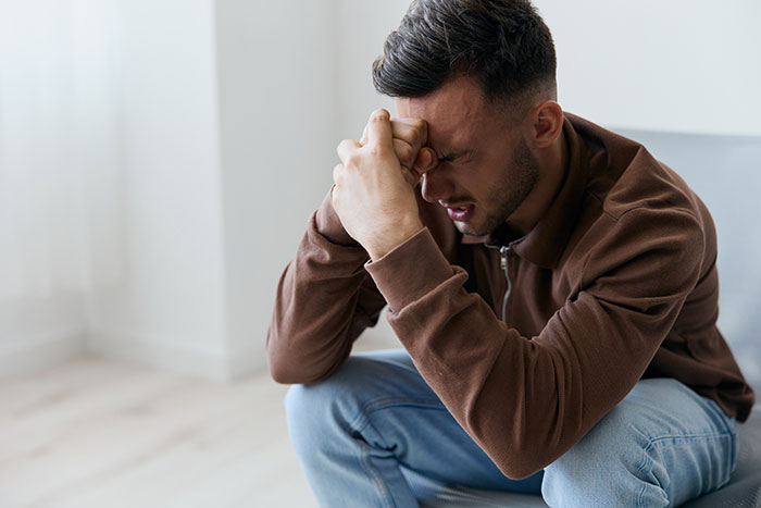 A man in emotional distress sitting on a couch, wearing a brown jacket and jeans.