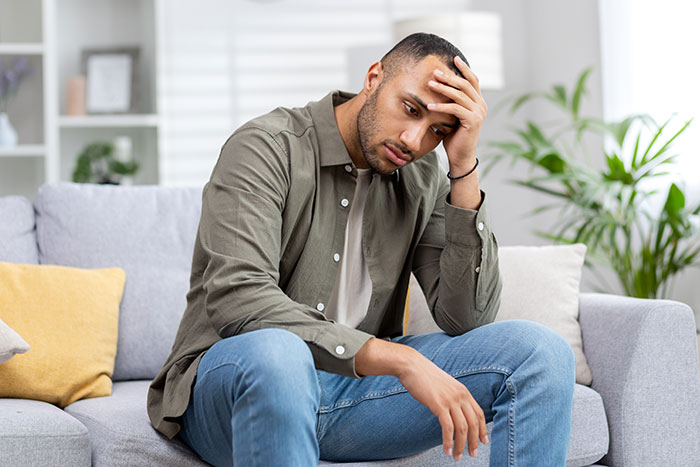 A man sitting on a couch with his hand on his forehead, looking distressed after a rejected proposal.