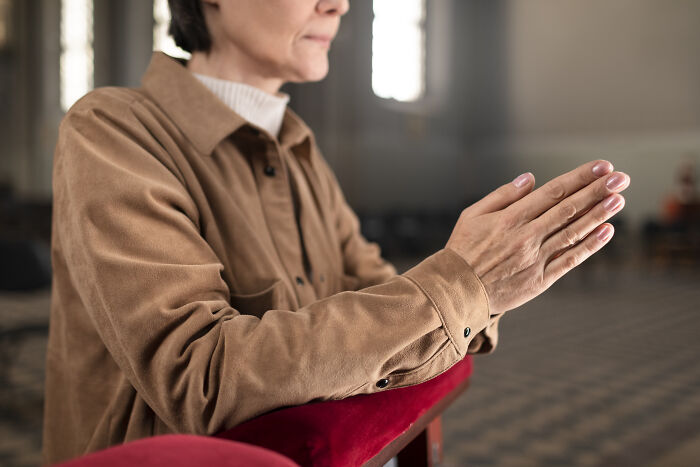 Person in a brown shirt praying with hands together in a dimly lit room, suggesting contemplation on human history scams.