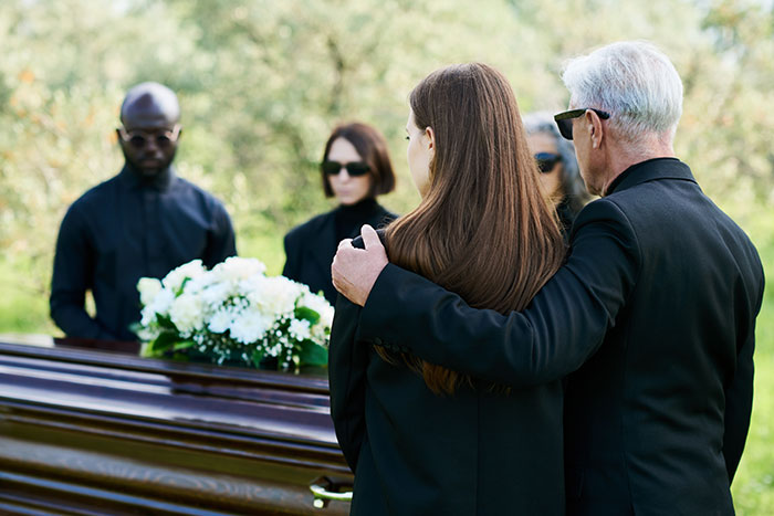 People in black attire at a funeral, standing by a flower-adorned casket outdoors.