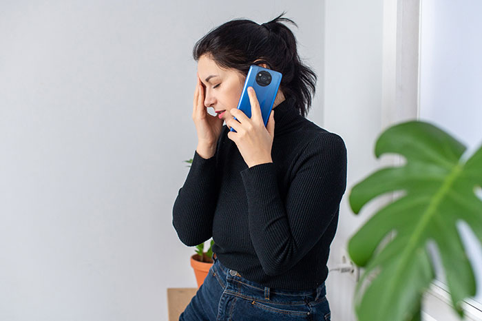 Woman in black sweater on phone, looking stressed near a plant.