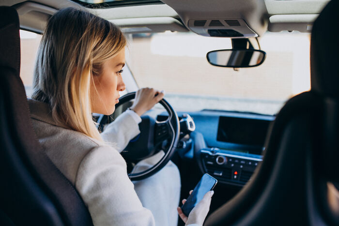 A woman driving a car, holding a smartphone, possibly distracted by the biggest scam.