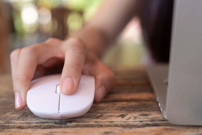 A hand using a white computer mouse on a wooden table next to a laptop, representing unethical life hack strategies.