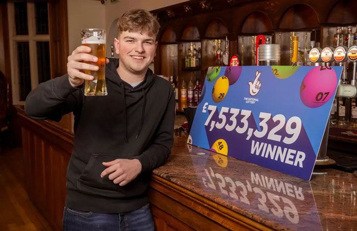 Man in a pub celebrating a £7.5M lottery win with a pint, standing next to a winning sign.