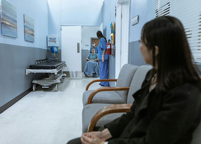 A woman in a hospital hallway watches nurses assist a patient in a wheelchair, highlighting American healthcare challenges.