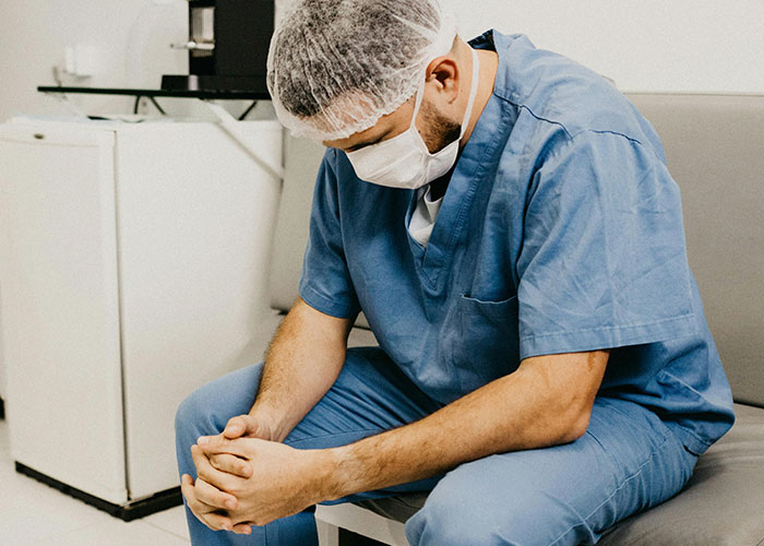 Healthcare worker in scrubs sitting on a bench, appearing stressed and contemplative in a hospital setting.