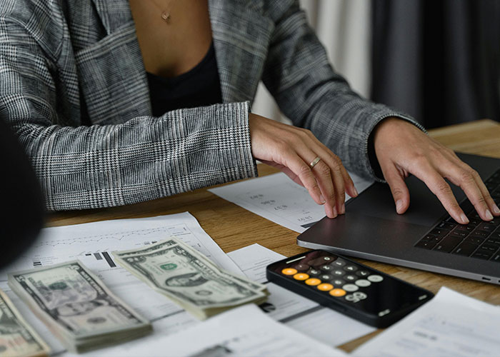 Person in plaid jacket using a laptop with cash and bills nearby, illustrating American healthcare costs.