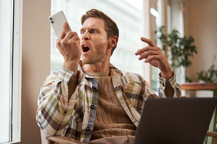 A frustrated person reacts to a spam call from a realtor, holding a smartphone and sitting near a window with a laptop.