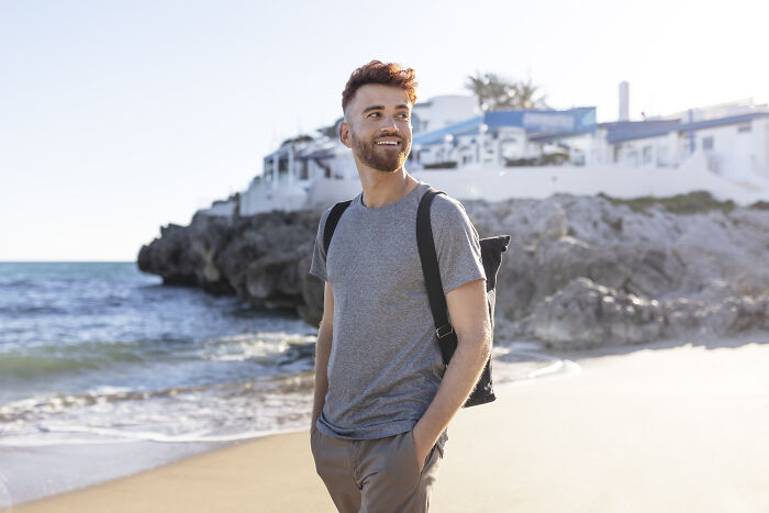 Man smiling on the beach, wearing a gray shirt and backpack, enjoying a getaway after a savage job exit.