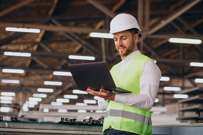 Construction worker in a warehouse wearing a hard hat and safety vest, using a laptop.