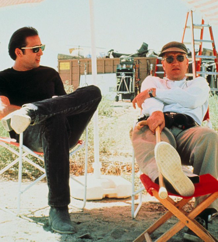 Two men sit in director's chairs on a film set, with one holding a cane and wearing sunglasses.