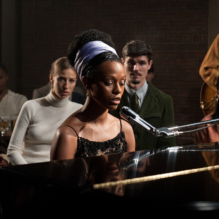 A woman with a headband sings at a piano, surrounded by an attentive audience.