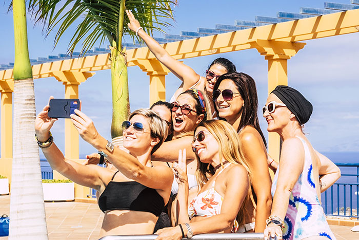 Group of women in swimsuits taking a selfie on vacation near palm trees, enjoying sunny weather.