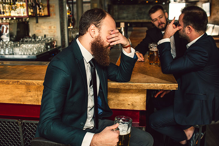 Three men in suits sitting at a bar, looking serious, with one covering his face; concept of homophobic confrontation.