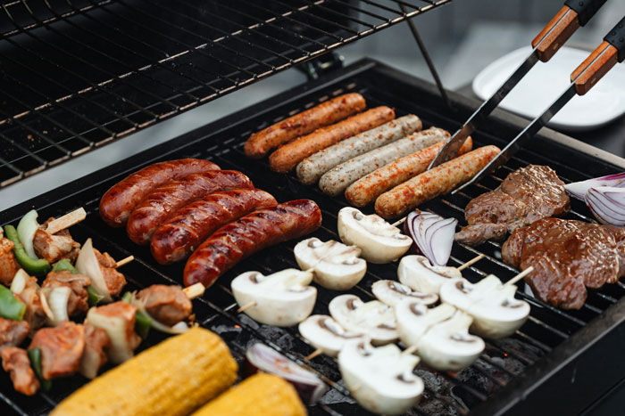 A family barbeque with sausages, corn, mushrooms, and onions on a grill.