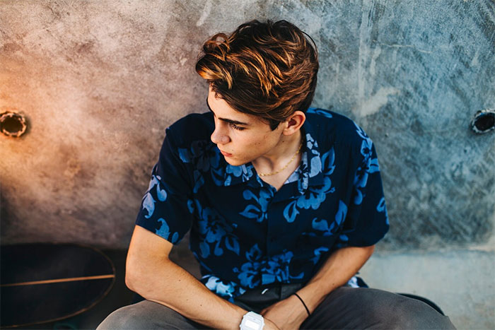 Young man in a blue floral shirt sitting against a concrete wall.