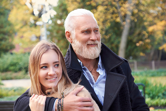 Older man and young woman smiling outdoors, showcasing age-gap relationships.