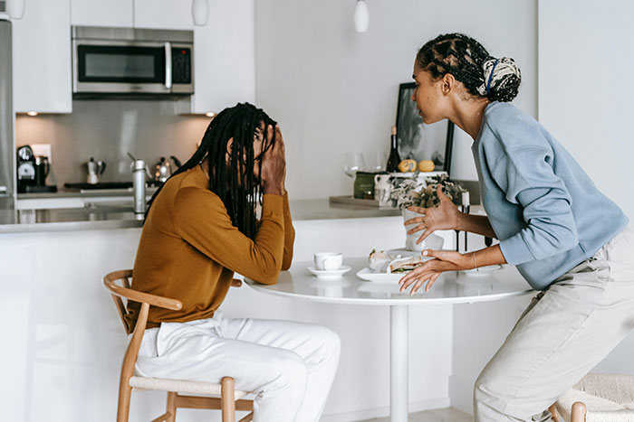 Couple having an argument at the kitchen table; man looks tired, covering his face, while woman gestures expressively.