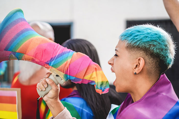 Person with blue hair and rainbow flag shouting through a megaphone covered in a rainbow cloth, highlighting hate crime awareness.