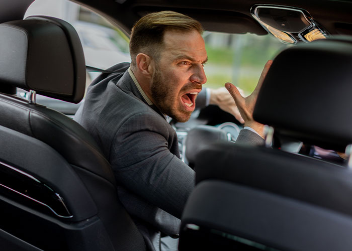 Hombre enojado en el asiento trasero de un coche, ilustrando una excusa increíblemente verdadera de estudiantes.