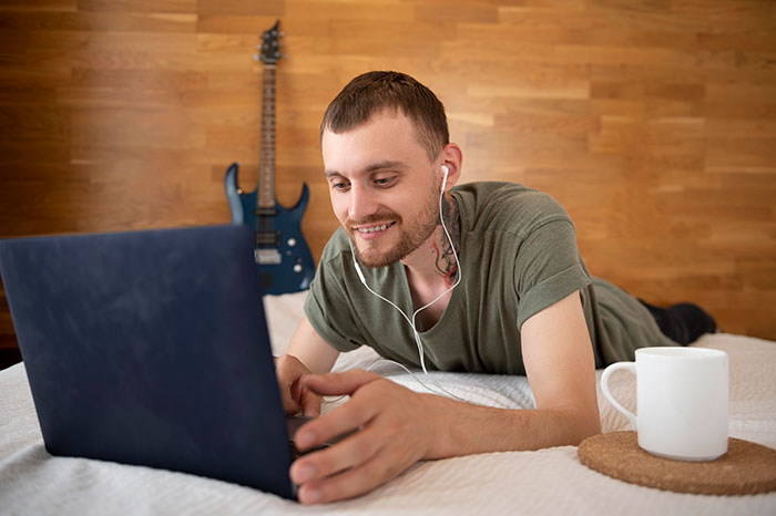 Man on bed using laptop with headphones, planning petty revenge.