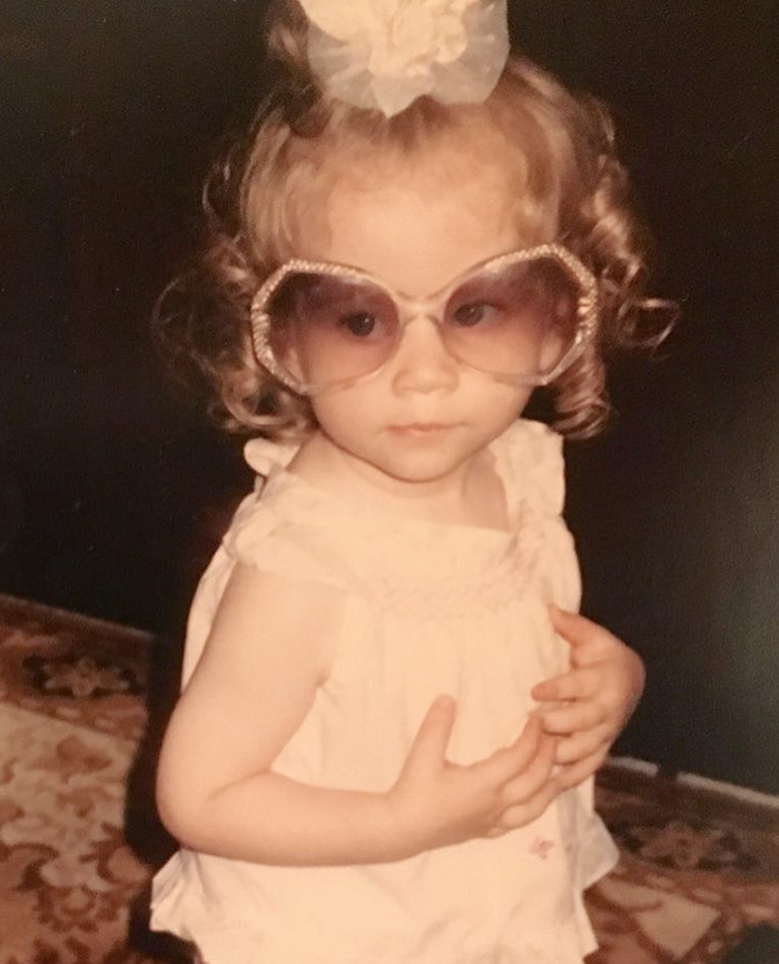 Young Michelle Trachtenberg with curly hair wearing oversized sunglasses and a dress.