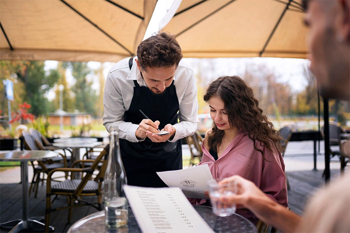 A bartender taking an order from a woman under a patio umbrella at an outdoor cafe.