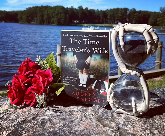 Romantic book by a lake with red roses and hourglass on a sunny day.