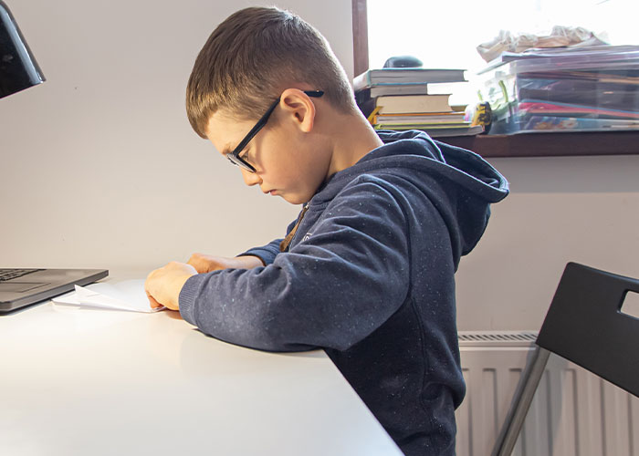 Niño con gafas en escritorio, escribiendo en papel bajo lámpara portátil, rodeado de libros y materiales escolares.