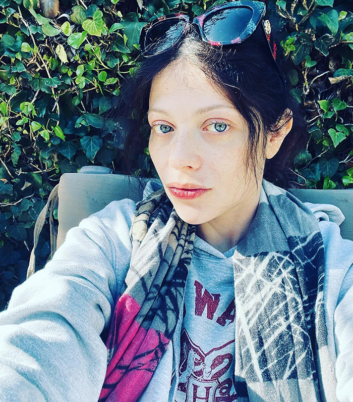 Michelle Trachtenberg wearing sunglasses on head, sitting in front of leafy background, in a casual hoodie and scarf.