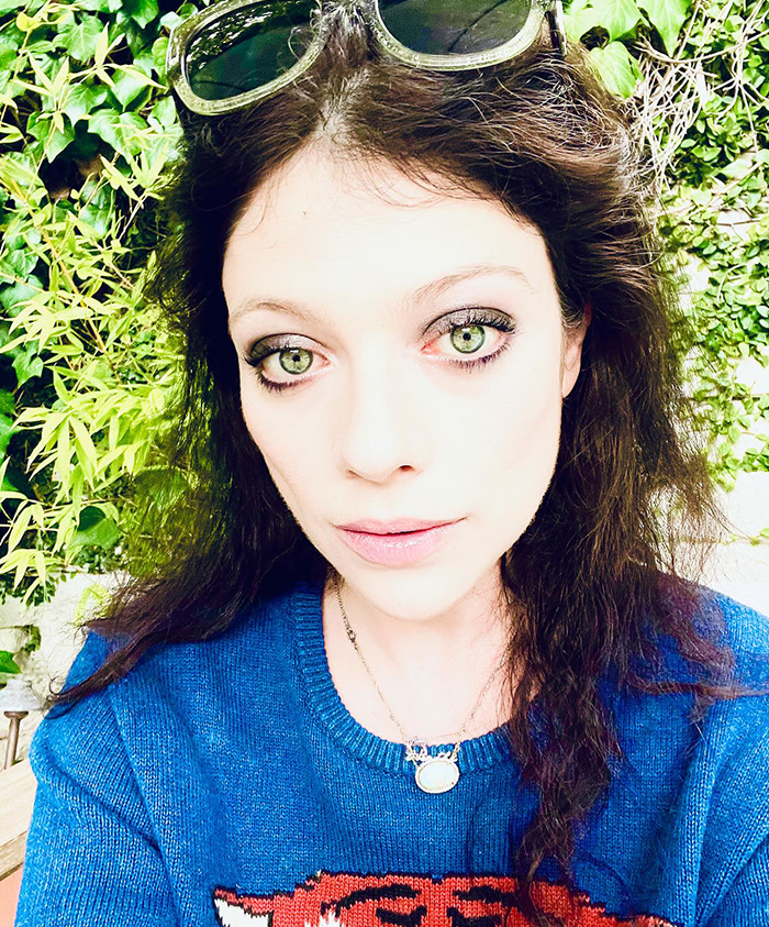 Michelle Trachtenberg in a blue sweater stands in front of green foliage, wearing sunglasses on their head.