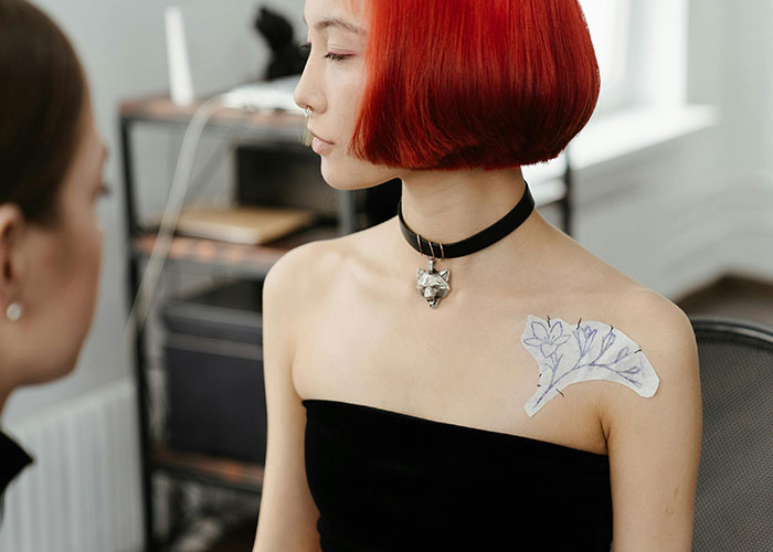 Person with red hair and choker showing a tattoo stencil on their shoulder, considering getting inked.