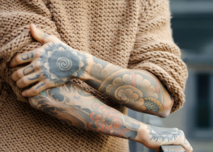 A person with floral and spiral tattoos on forearms, wearing a knitted sweater, illustrating things to know before getting inked.