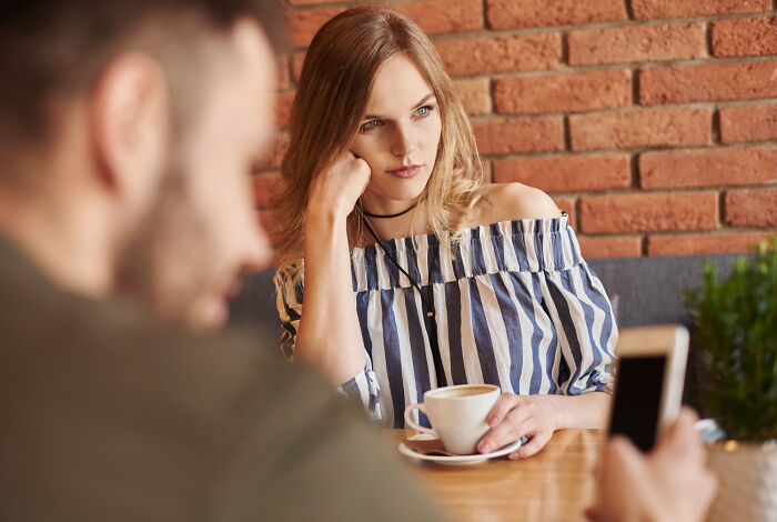 Woman bored on a date, resting her chin in her hand, holding a coffee, while the man looks at his phone.