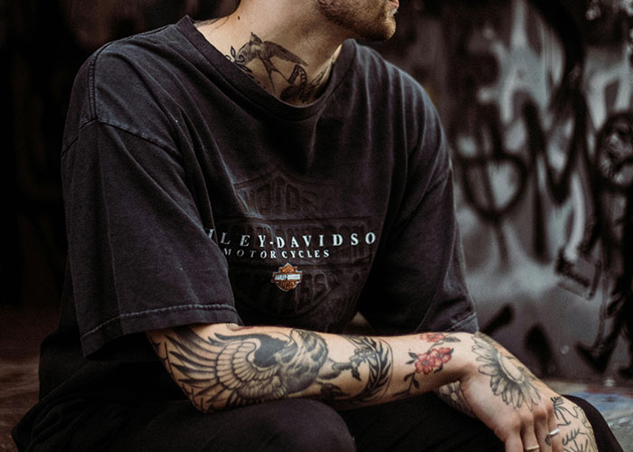 Person with tattoos wearing a Harley-Davidson shirt, sitting against a graffiti wall.