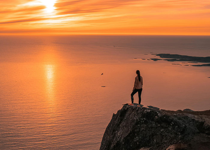 Person stands on a cliff overlooking the ocean at sunset, surrounded by wholesome, warm orange hues.