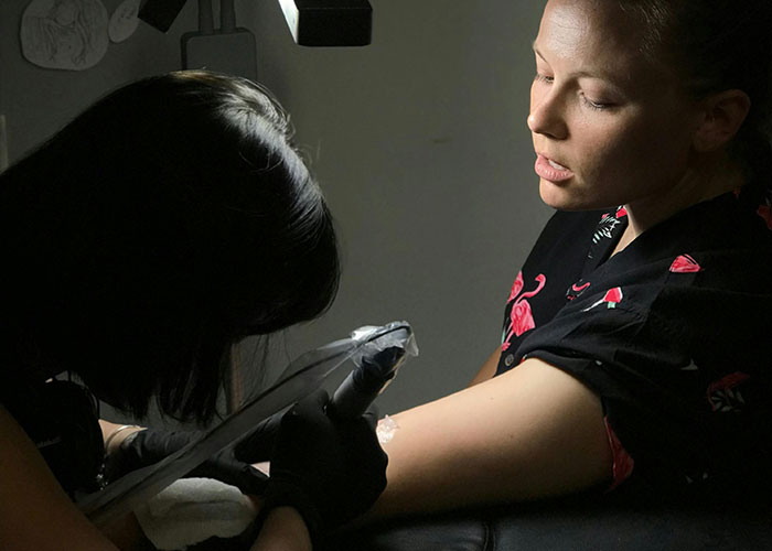 A person getting inked, while an artist focuses on tattooing their arm in a dimly lit setting.