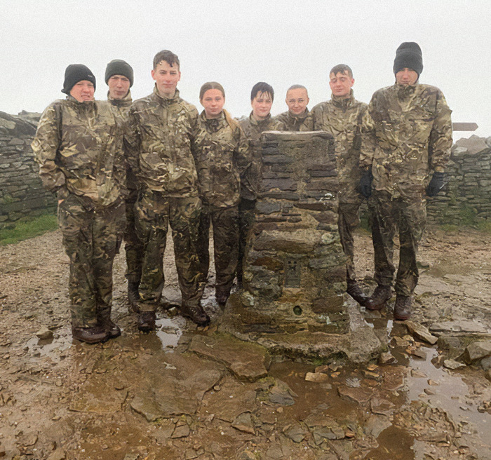 Soldiers in camouflage stand outdoors by a stone marker on a misty day at a base.