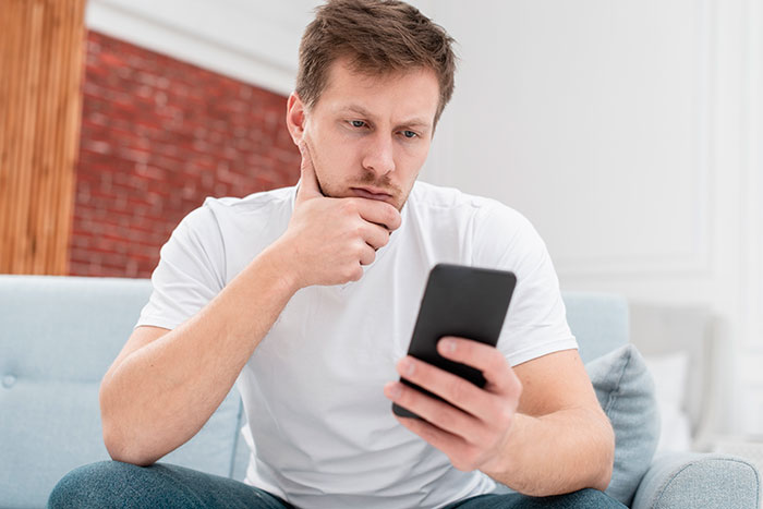 Man in white shirt on couch looking at phone, pondering family responsibilities.