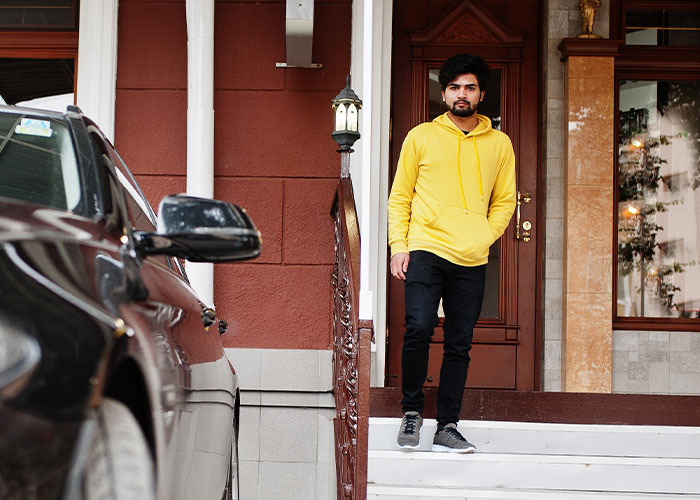 Man in a yellow hoodie on a porch looks at a car parked in a neighbor's driveway.