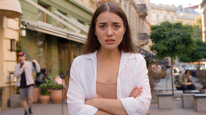 Young woman on city street, looking concerned, crossing her arms, related to dad’s girlfriend parenting dynamic.