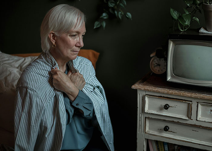 Elderly woman with dementia sitting in a softly lit room, wearing a striped shirt, near a vintage TV.