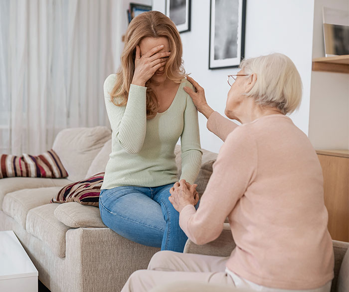 Woman distressed about inheritance news while elderly mother comforts her on a sofa.