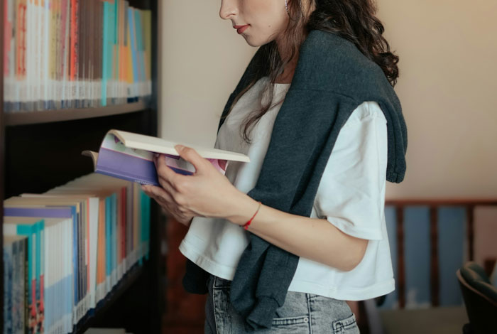 Woman reading in a library, wearing a white shirt and jeans, showcasing her pursuit of an engineering degree.